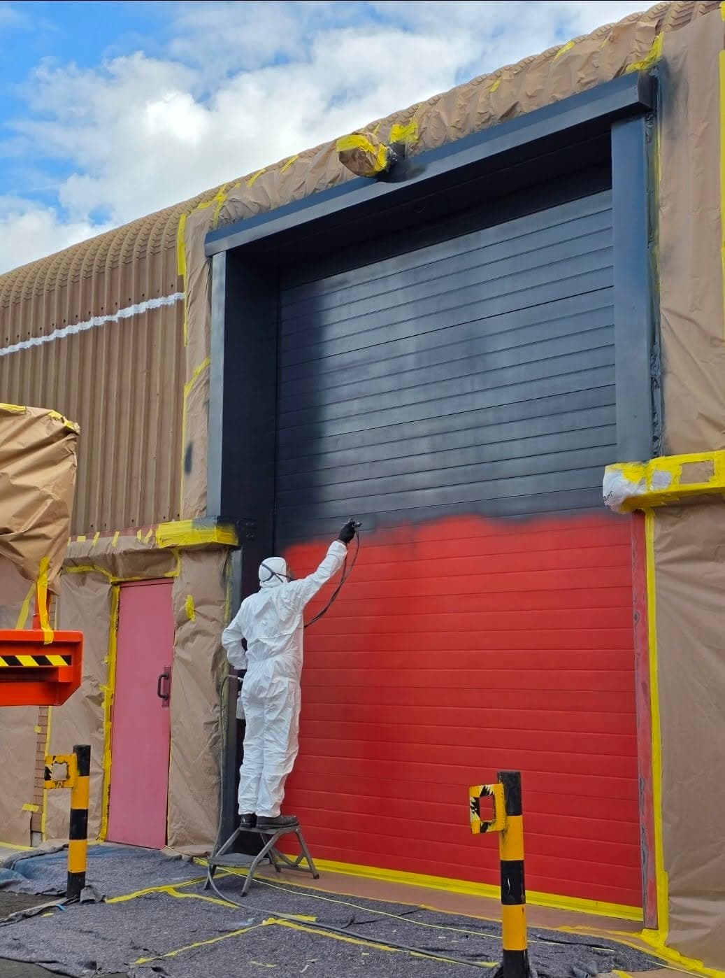 Sprayer in protective overalls respraying a red roller shutter dark grey, with the surrounding cladding masked
